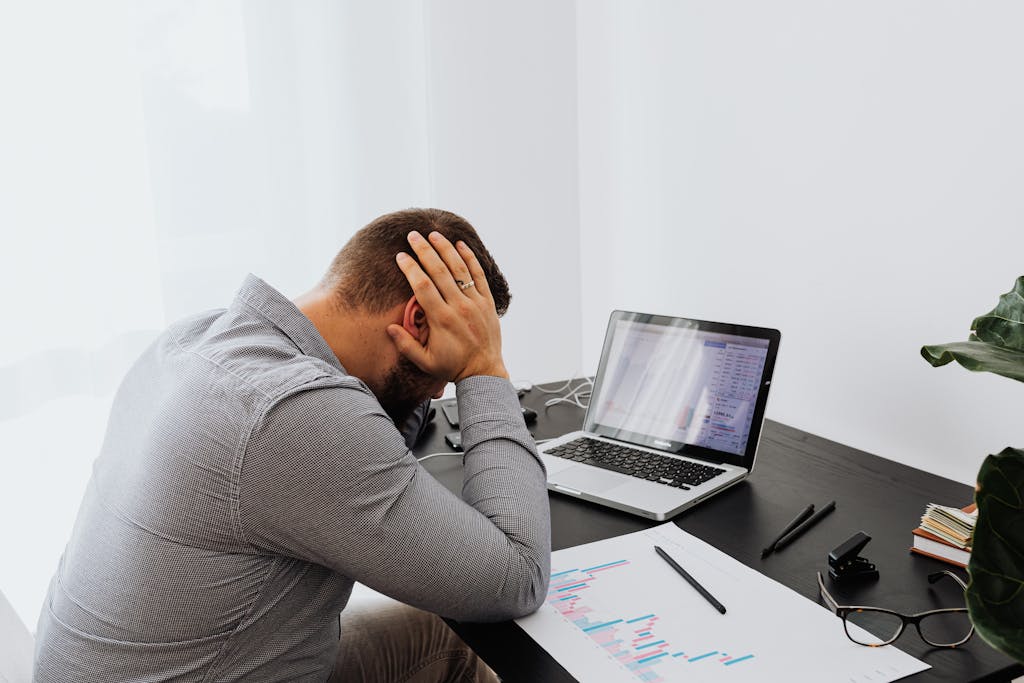 A man looking stressed at his desk, hands on head, with a laptop and graphs.
